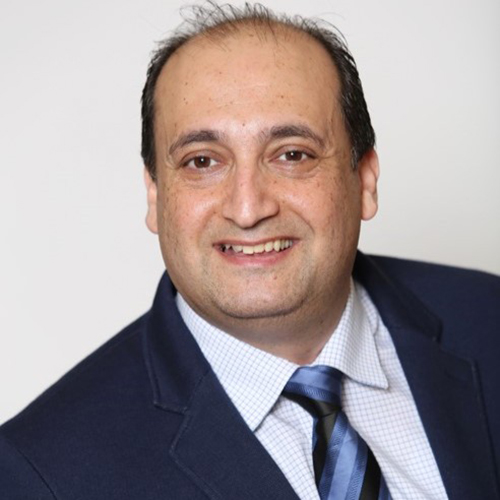 A portrait of new DIA Global Chief Executive Marwan Fathallah smiling in a dark navy blue suit and white patterned-style button-up dress shirt underneath held together by a multi-colored (blue and black) tie