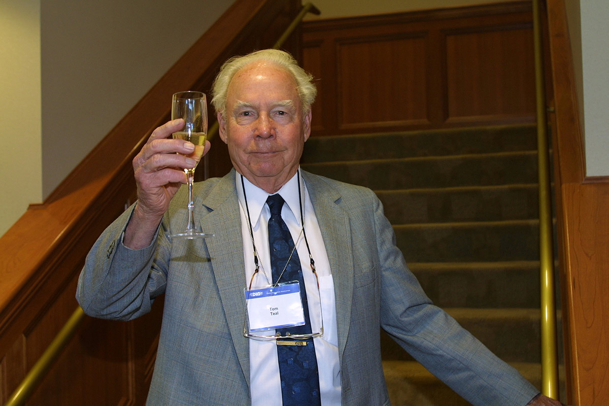 Close-up indoor photograph perspective of Tom Teal in a grey blazer suit and white closed button-up dress shirt with a dark blue tie as he holds a glass cup with champagne inside the glass while Tom stands on a staircase step with his left arm on the guardrail at the DIA headquarters