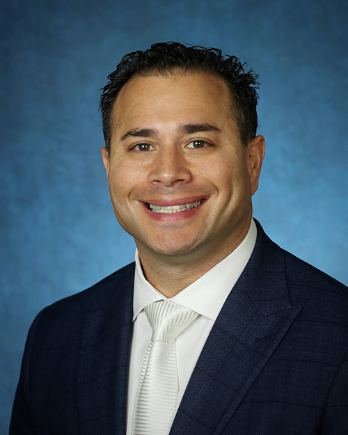 Portrait orientation close-up headshot photograph of Miguel Romero smiling in a dark navy blue business blazer suit and white button-up dress shirt underneath and white striped pattern style tie equipped
