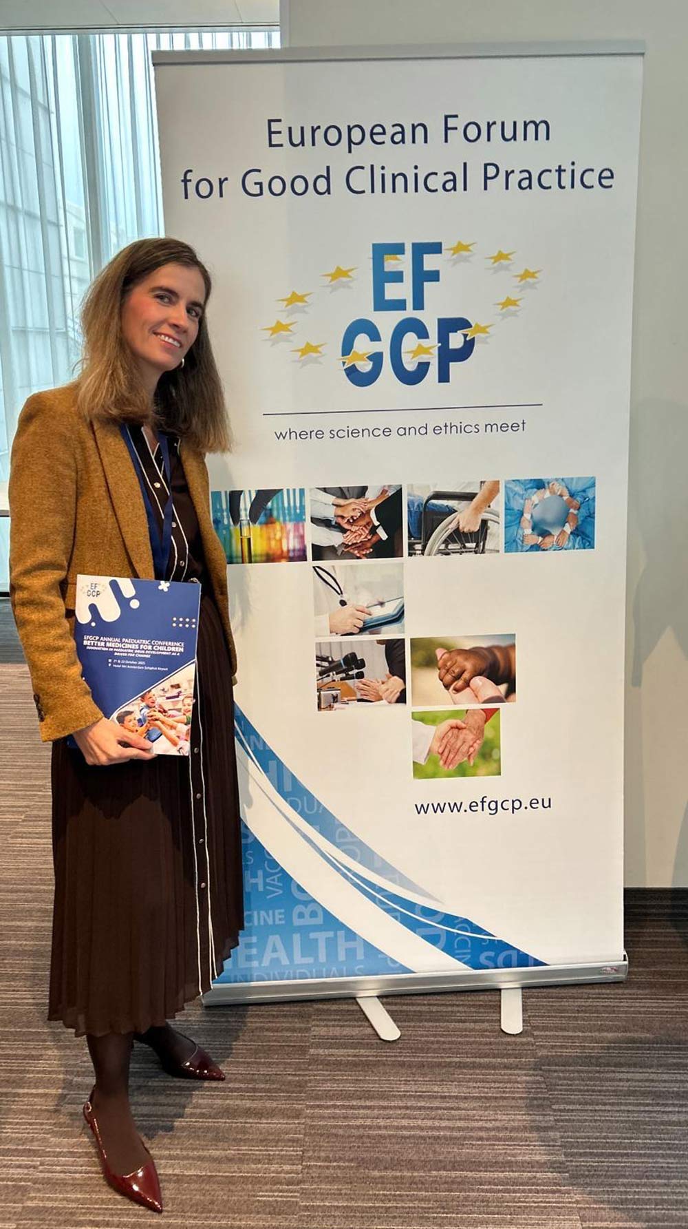 A woman stands next to a large "European Forum for Good Clinical Practice" banner holding a conference program.