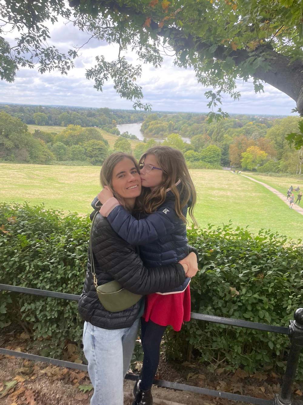 A woman and a young girl in a blue puffer jacket embrace while standing on a hill overlooking a scenic river valley.